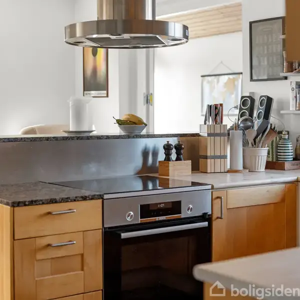 Komfur står stille i moderne køkken med træskabe, sort bordplade og emhætte. Køkkenredskaber og krydderier er organiseret på bordet og hylderne. Vindue viser udsigt til tagetage.