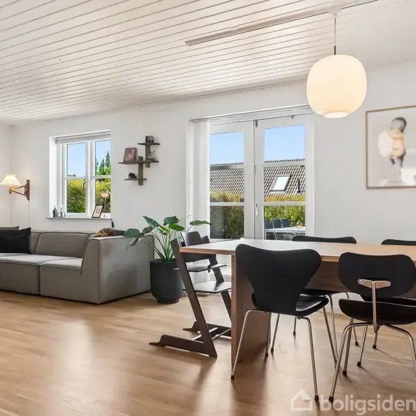 En stue med en grå sofa i hjørnet. Et spisebord med sorte stole står i midten. Værelset har trægulv, hvide vægge, og store vinduer med udsigt til have.