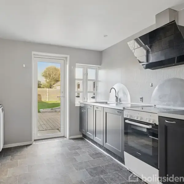 Et moderne køkken med sort-hvide skabe står i et lyst rum. En dør fører ud til en have, og en komfur og emhætte ses tydeligt.