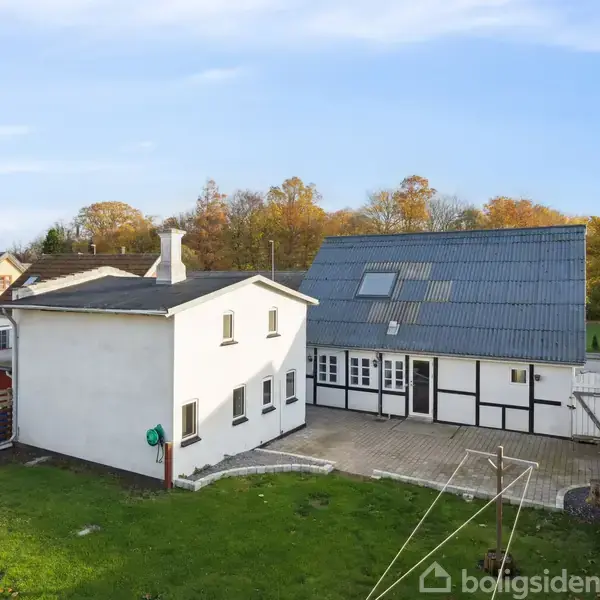 Et hvidt hus med sorte detaljer står midt i en grøn have. Der er en terrasse foran huset. Baggrunden har træer med efterårsfarver og flere nabohuse i nærheden.