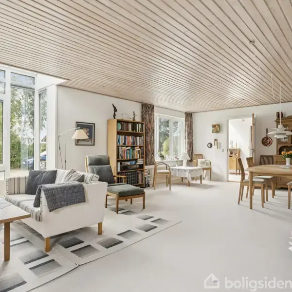 Stue med træloft og store vinduer. Møbleret med sofaer, bogreol og spisebord. Blid belysning og tæpper på gulvet. Udenfor ses haveelementer gennem vinduerne.