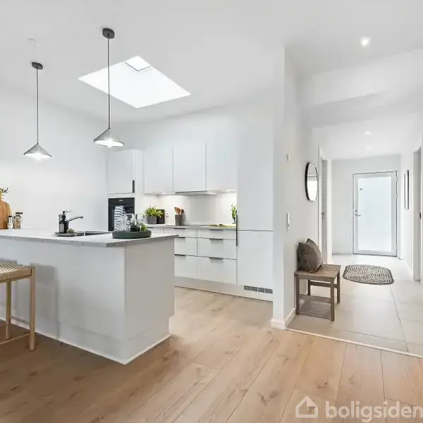Et moderne køkken med hvidt interiør har et langt køkkenbord med to barstole. Loftslamper hænger over bordet, og gangen i baggrunden fører til en lys indgang.
