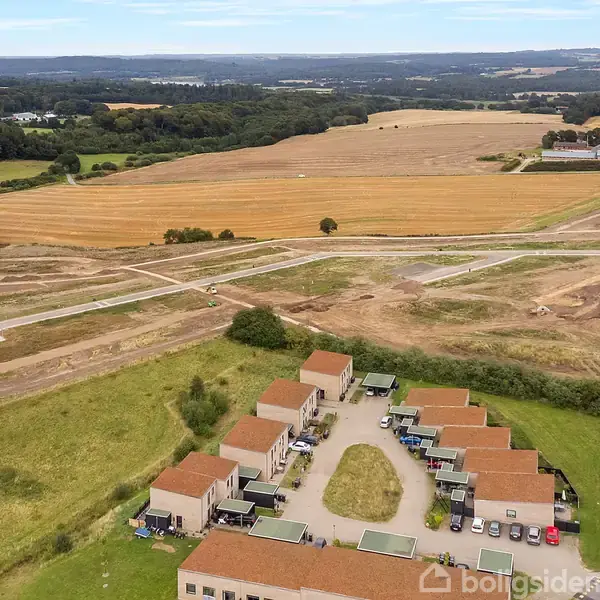 Rækkehuse med røde tage står i række, omgivet af grønne marker og en byggeplads med veje under konstruktion. I baggrunden ses et stort åbent landskab med marker og skove.