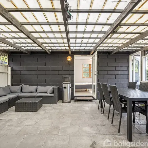 Udendørs overdækket terrasse med en sort sofa og bord til venstre, samt et langt spisebord med flere stole til højre. Baggrund af sorte teglsten på væggen.