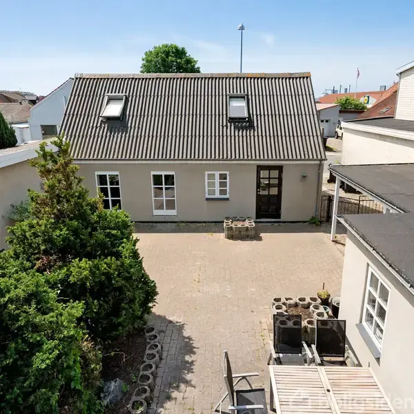 Et hus med skråt tag og loftsvinduer står foran en flisebelagt gårdsplads omgivet af buske og bygninger. En stak blokke og træmaterialer ligger på gårdspladsen.