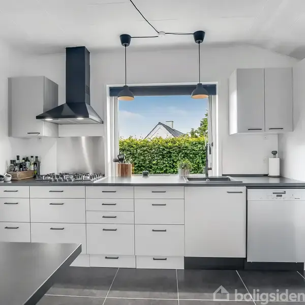 Et moderne køkken med hvide skabe og sorte bordplader. To lamper hænger over en indbygget kogeplade. Et vindue viser en hæk og hus i baggrunden.