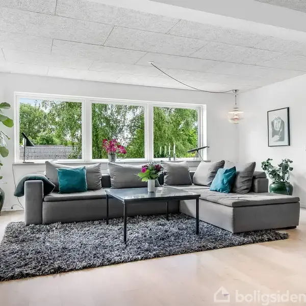 En grå hjørnesofa står på et mørkt tæppe i en lys stue. Sofaen er pyntet med farverige puder, og et sofabord med blomster står foran. Store vinduer viser grønne træer udenfor.