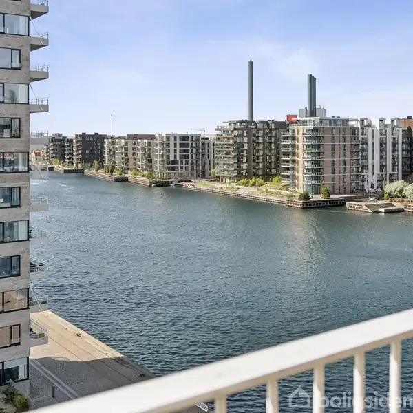 Udsigt fra en altan over en bred kanal omkranset af moderne lejlighedsbygninger med altaner. Bygningerne har flere etager og står langs vandet under en klar blå himmel.