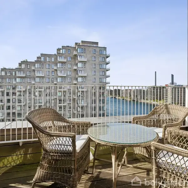 Fletstole og et glasbord står på en balkon med udsigt til moderne lejlighedskomplekser og en flod. Klar blå himmel.