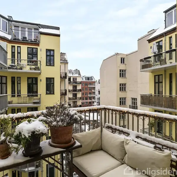 En altan med snebelagte planter og lysdekorationer. Beliggende mellem gule lejlighedsbygninger i en bymæssig baggård. Flere etager kan ses med balkoner og vinduer.