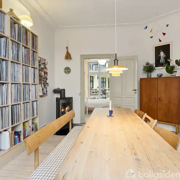 Langt træbord med bænke står centralt i et lyst rum. Til venstre er en stor reol fyldt med plader. I baggrunden ses en brændeovn og dør til altan.