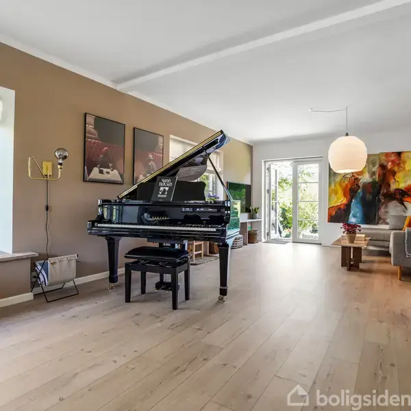 Flygel placeret i en stue, hvor det står ved siden af en væg med kunst på. I baggrunden ses en sofa og et bord, med store vinduer mod haven.