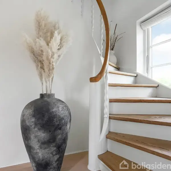 En stor, sort vase med tørrede pampasgræs står stille ved en hvid, snoet trappe med trætrin. Et vindue i baggrunden slipper sollys ind i rummets lyse atmosfære.