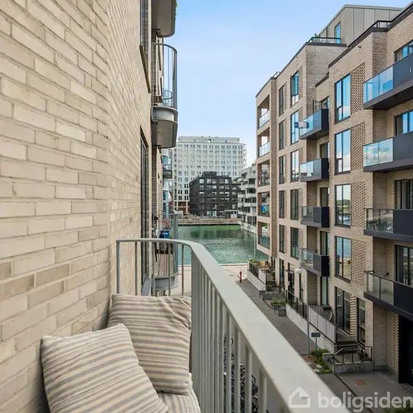 En balkon med lyse hynder på en bænk, der vender mod en moderne lejlighedsbygning. I baggrunden ses flere bygninger langs en smal vandkanal.