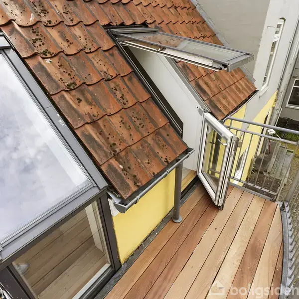 En balkon med trædæk strækker sig ud fra en taglejlighed. Taget er dækket af røde teglsten. På væggen er der to vinduer, et åbent. Omgivet af andre bygninger.