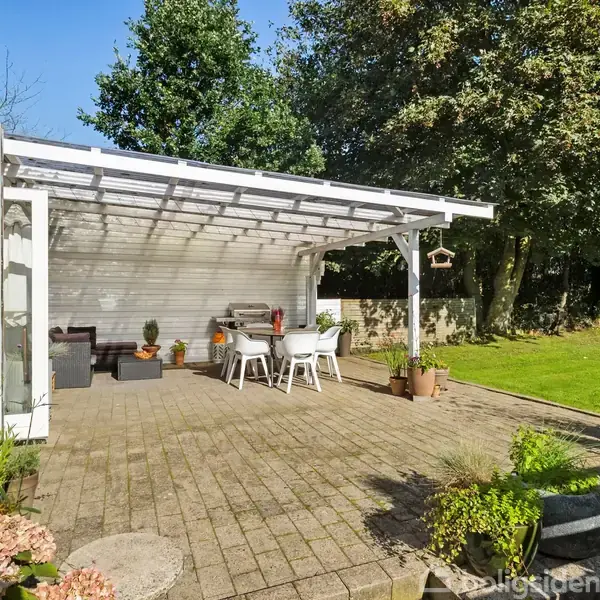 Terrasse med overhængende tag af hvidt træ, møbleret med spisebord og stole. Omgivet af planter og buske, grænser op til en grøn græsplæne og træer i baggrunden.