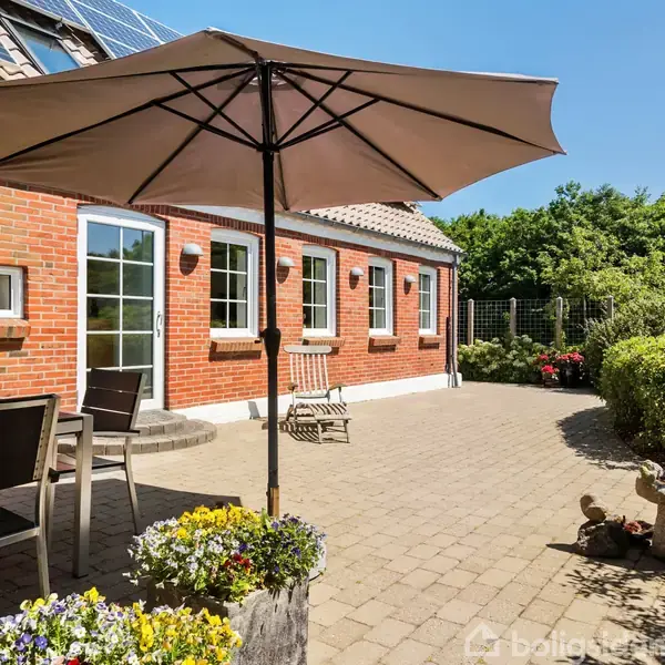 En terrasse med flisebelægning har en parasol i midten, omgivet af grå havemøbler. I baggrunden ses et rødt murstenshus med flere vinduer og grøn beplantning.