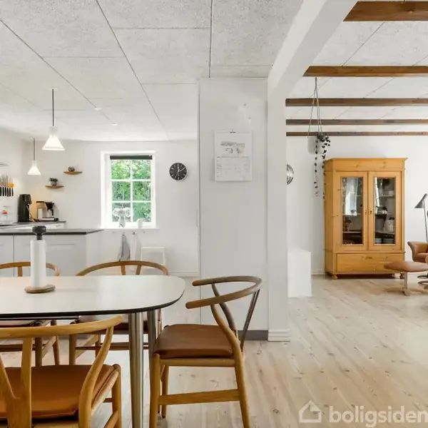 Spisebord står stille i et lyst, åbent køkken-alrumsområde. Omkring er der moderne hvide køkkenelementer, træstole, og hængelamper. I baggrunden ses en stue med træmøbler og store vinduer.
