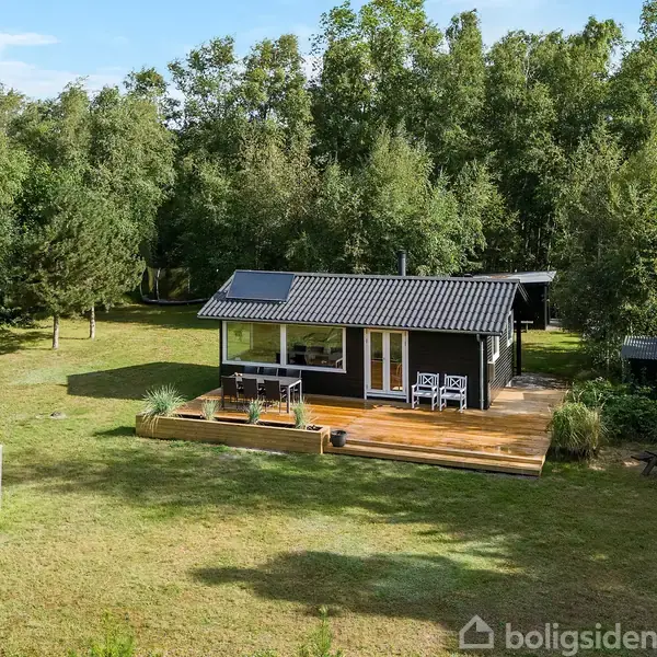 Sommerhus med sort facade står stille på en græsplæne omgivet af træer. En træterrasse med havemøbler er foran huset, og et lille fodboldmål ses på plænen.