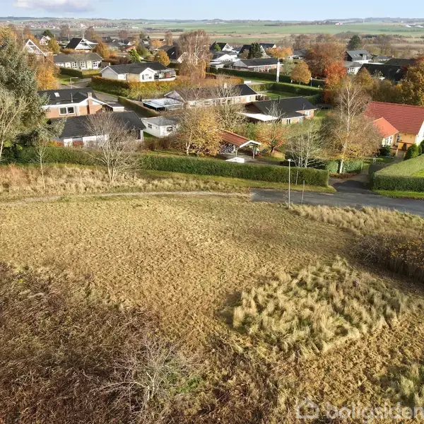 Græsklædt område ligger brak, omgivet af parcelhuskvarter med træer og hække i efterårsfarver. Rundkørsel og marker ses i baggrunden.