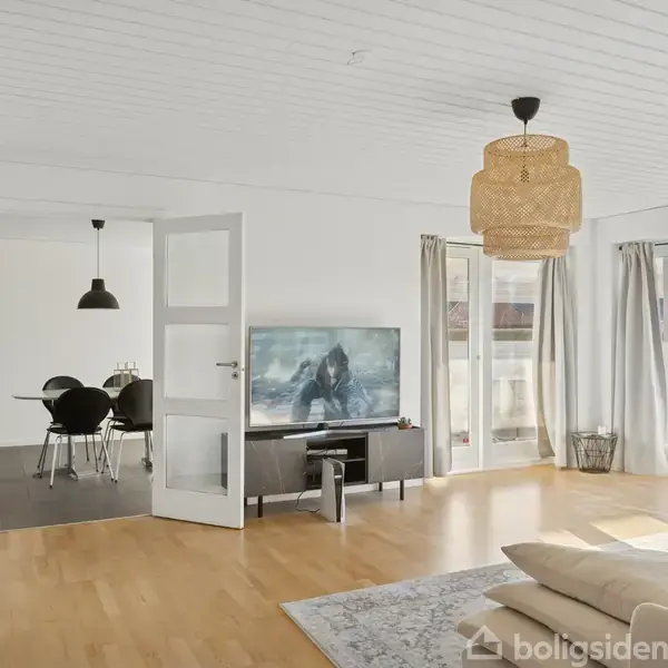 En stue med lyse trægulve og store vinduer, der lader lys strømme ind. Et TV står på et lavt møbel, mens stuen grænser op til en spisestue med spisebord.