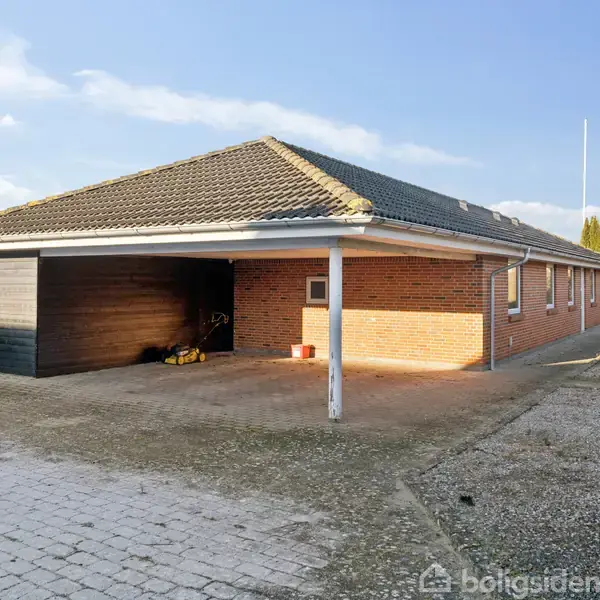 En rød murstensbungalow med tag af mørke fliser har en integreret carport til venstre. Omgivet af grusbelagt indkørsel og hække under en blå himmel.