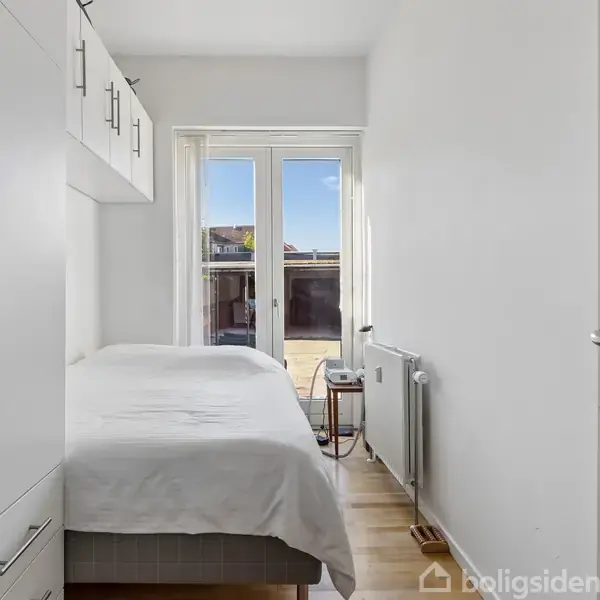 Seng med hvidt sengetøj står i et lyst rum med trægulv. Over sengen er der indbyggede skabe. To glasdøre leder ud til en terrasse. Radiator ved døren.