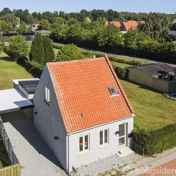 Et lille hus med rødt tegltag står i en frodig have med græsplæne og træer. En carport er ved siden af huset, omgivet af grønne hække og nabohuse.