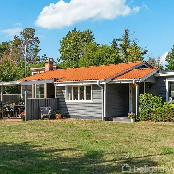 Et gråt sommerhus med rødt tag står på en græsplæne. Huset har en terrasse med havemøbler. Omkring huset er der træer og buske under en blå himmel med skyer.