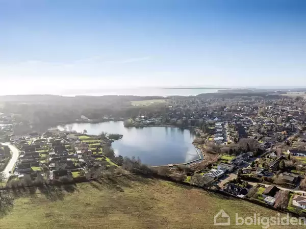 En sø i centrum omgives af huse og grønne områder, indrammet af en klar, blå himmel. Byen spreder sig ud i et bakket, skovklædt landskab.