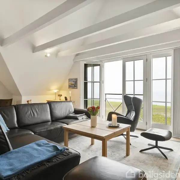 Stue med sort lædersofa og lænestol omkring et træbord, placeret ved store glasdøre. Udenfor ses græsplæne og himmel. Kunstmalerier pryder væggene, og rummet har synlige loftsbjælker.