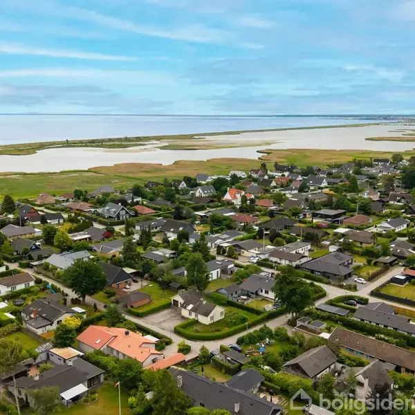 Beboelsesområde præsenterer en samling af huse omkranset af frodigt grønt, nær åbne marker og en bred vandstrækning i baggrunden, under en klar, blå himmel.