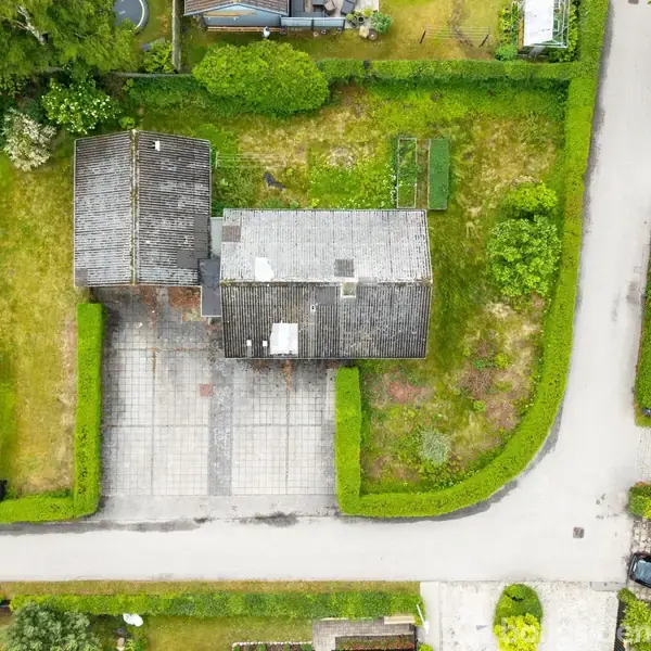 Et hus med to sammenhængende bygninger i en grøn have. Omgivet af en hæk og placeret ved en vej med fortove. To biler ses parkeret på en tilstødende grund.