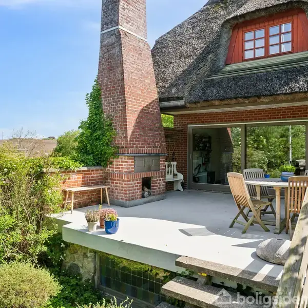 En terrasse med bord og stole står opstillet ved en murstensskorsten. Den er omgivet af grønne buske og planter, med en glasdør der fører ind i et stråtækt hus.