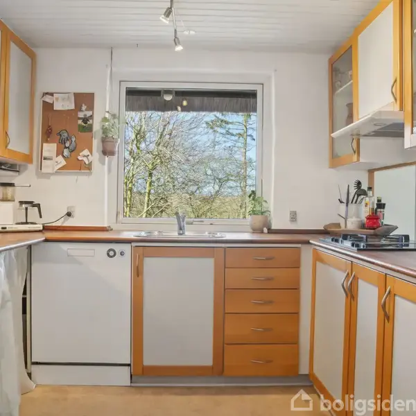 Køkken med hvide og træfarvede skabe, placerer køkkenmaskiner langs bordpladen. Vindue med udsigt til trætoppene i baggrunden. Knagerække og opslagstavle på væggen.