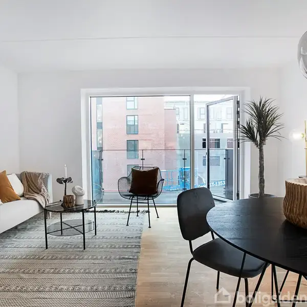 En stue med sofa og sofabord i venstre del, spisebord med stole til højre. Lysindfald fra en stor altandør fører til en udsigt over bygninger udenfor.