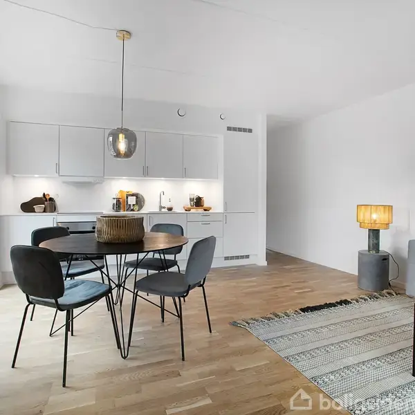 Et moderne køkkenalrum med spisebord, omgivet af fire stole. Køkkenet i baggrunden har hvide skabe og opvaskemaskine. Til højre står en grå sofa ved siden af en lille lampe på et tæppe.