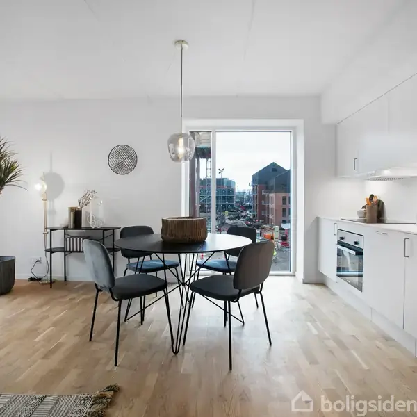 Et lyst køkken-alrum med et sort rundt spisebord og fire stole i midten. Til venstre står en plante, og til højre er der en hvid køkkensektion med indbygget ovn.