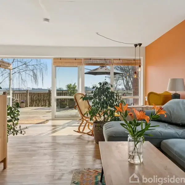 Stue med gråt sofamøbel og træmøbler, omgivet af indendørs planter og orange væg. Store vinduer åbner ud mod en terrasse med udsigt til have og træer.