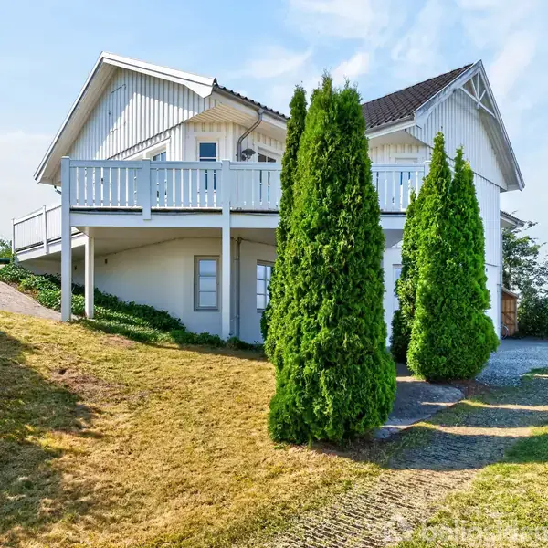 Hvide hus med veranda, omgivet af grønne buske i forgrunden. Plænen er let skrånende mod huset. Klar blå himmel i baggrunden.