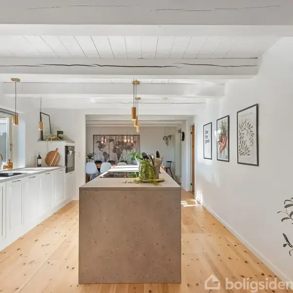 Et moderne køkken med en stor, grå køkkenø, omgivet af hvide skabe og træpaneler. Køkkenet har ophængte lamper, træloftsbjælker og vægdekorationer, der skaber en lys og stilfuld atmosfære.