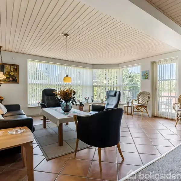 En stue med sorte møbler og blomsterdekoration på bordet. Rummet er lyst med store vinduer og gulvlamper. To fletstole er placeret nær vinduet, omgivet af billeder på væggene.