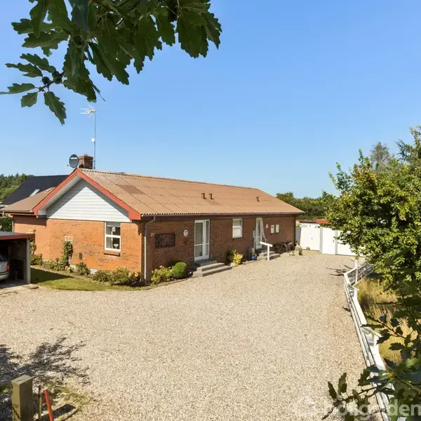 En rød murstenshus med garage til venstre, omgivet af en grusindkørsel og grønne buske, under en blå himmel.