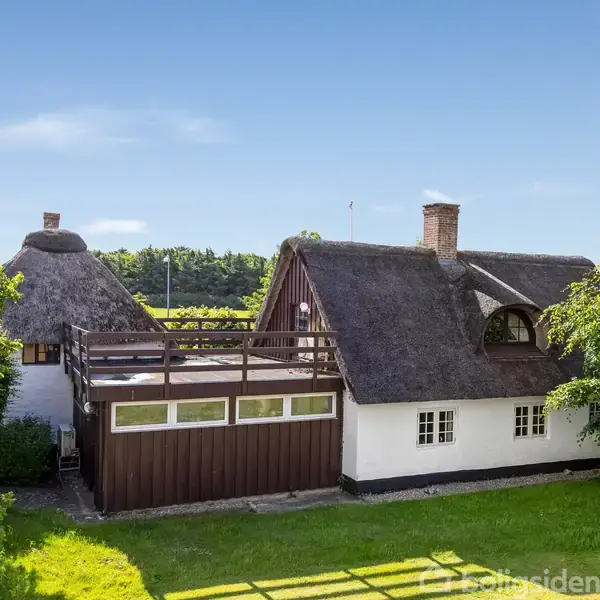 Stråtækket hus med hvide vægge og træterrasse ligger omgivet af grønne træer og græsplæne. Huset har en skorsten, og terrassen er aflang med trægelænder.