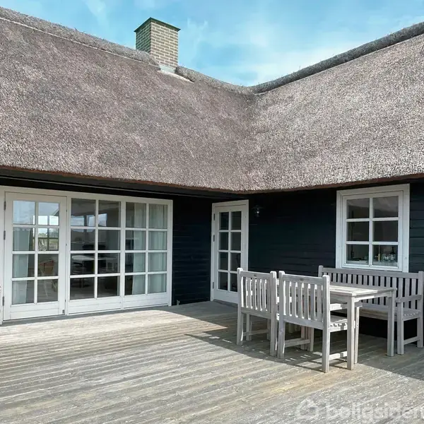 Stråtækt hus med mørk facade og store glasdøre. Træmøbler står arrangeret på en terrasse af træplanker. Himlen er blå og bygningen omgivet af rolig natur.