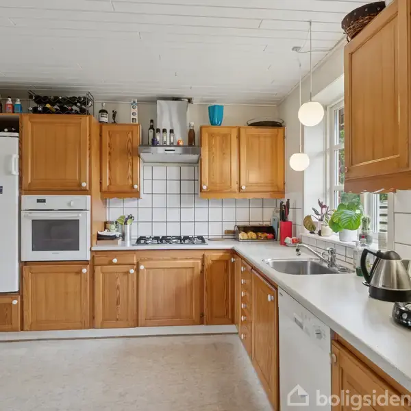 Køkken med træskabe, kogeplade og indbygget ovn. Kedel og kaffemaskine står på bordet. Køleskab med magneter og vindue med planter i en lys indretning.