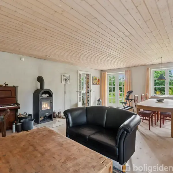 Sort lædersofa står i en stue med trægulv og loft, ved siden af et træbord. Brændeovn og klaver på venstre side, spisebord og store vinduer i baggrunden.