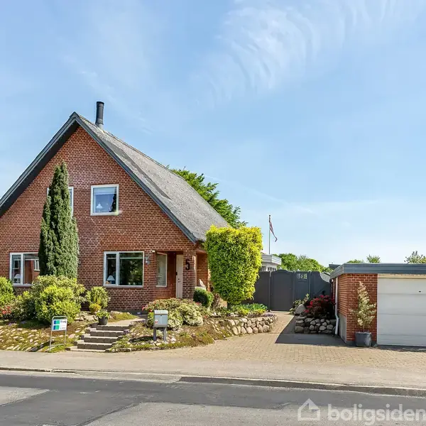 Rødt murstenshus med stejl gavl står stille i en velplejet have med forskellige buske. En garage findes til højre, og himlen er klar og blå.