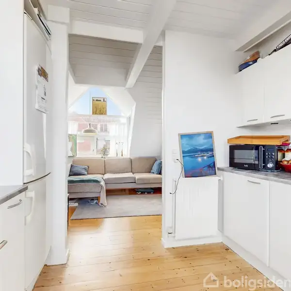 Et hvidt køkken i brug, oven på brunt trægulv, med mikroovn og frugt på bordet. I baggrunden ses en lys stue med sofa nær et stort vindue.