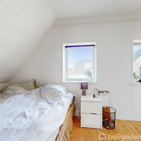 Seng står i et lyst loftværelse ved siden af et natbord med lampe og bøger. To små vinduer lader naturligt lys ind. Gulvet er i lyst træ.